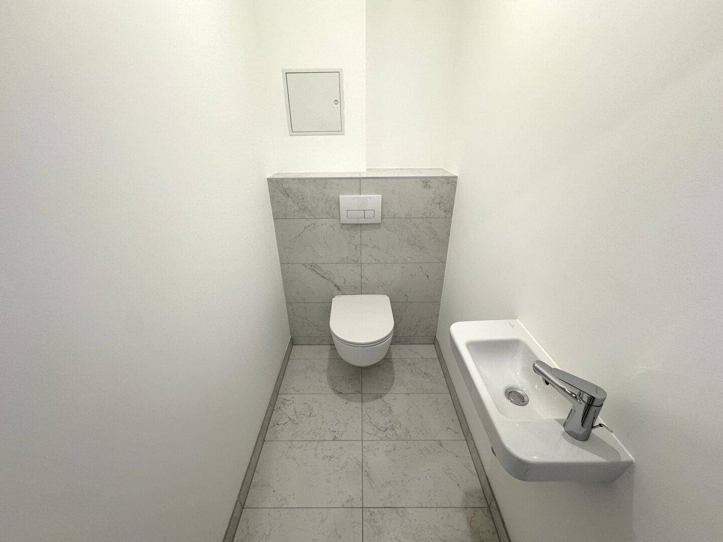 Modernes Gäste-WC mit wandhängender Toilette, kleinem Waschbecken und grauen Fliesen an der Wand.