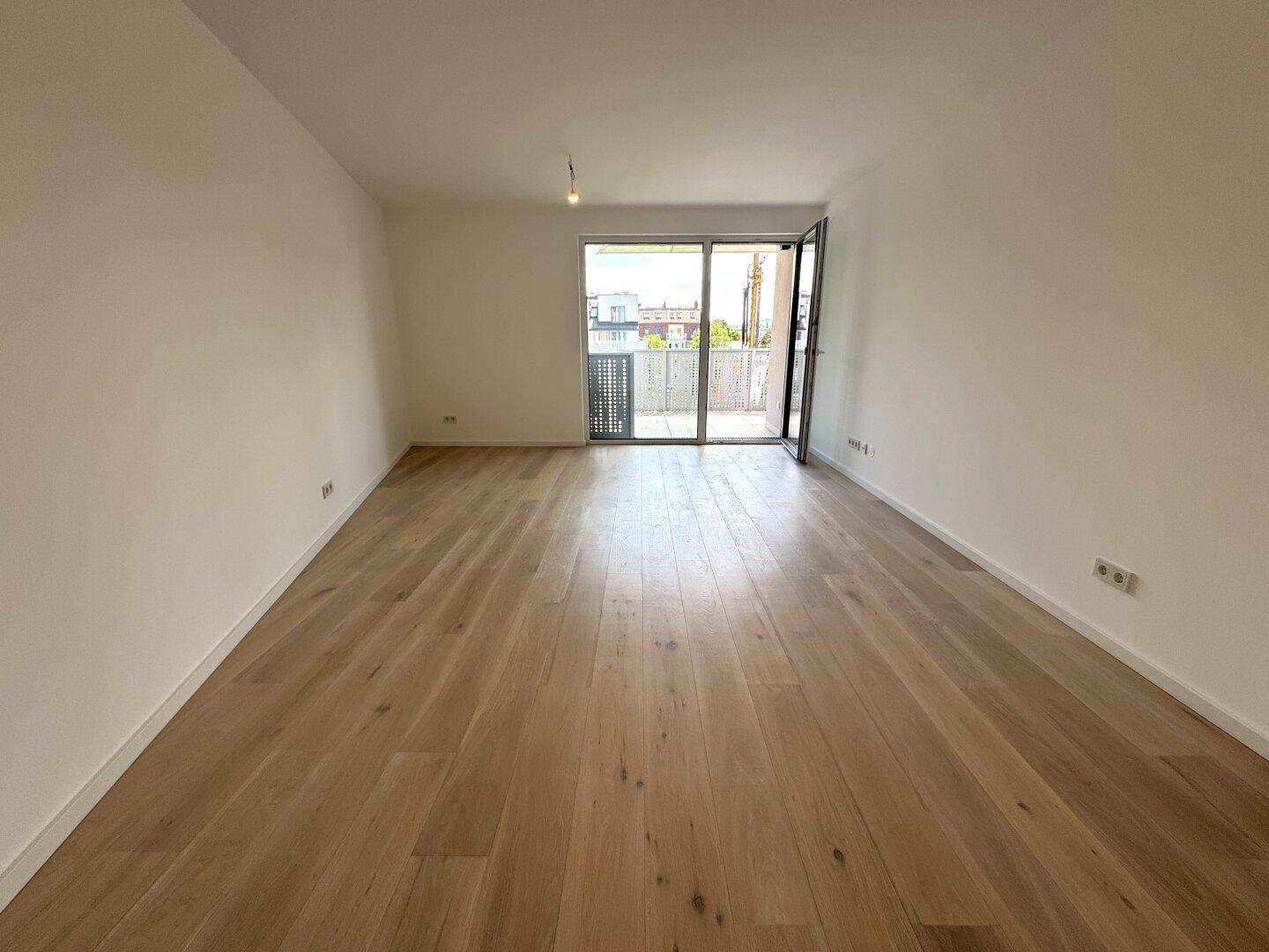 Helles, leeres Wohnzimmer mit Holzboden und Zugang zu einem Balkon.
