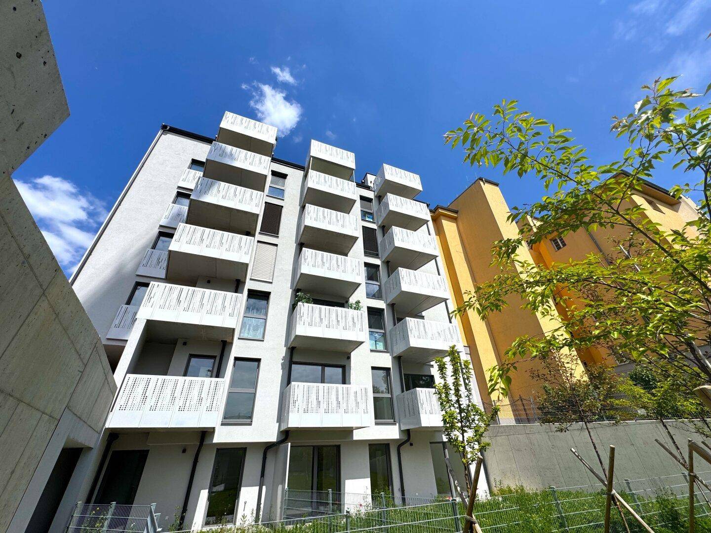 Aufwärtsblick auf die weiße Fassade des modernen Apartmentgebäudes mit zahlreichen Balkonen und klarem Himmel.