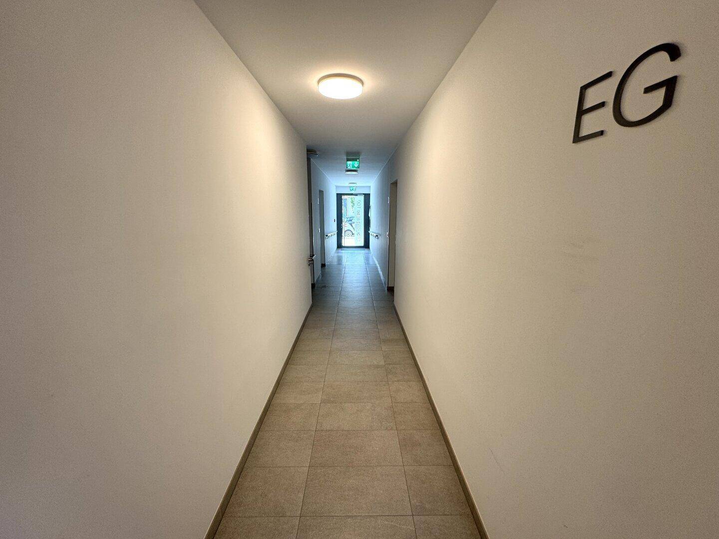 Langer, heller Flur mit beigefarbenen Fliesen und weißen Wänden, der zum Ausgang führt.