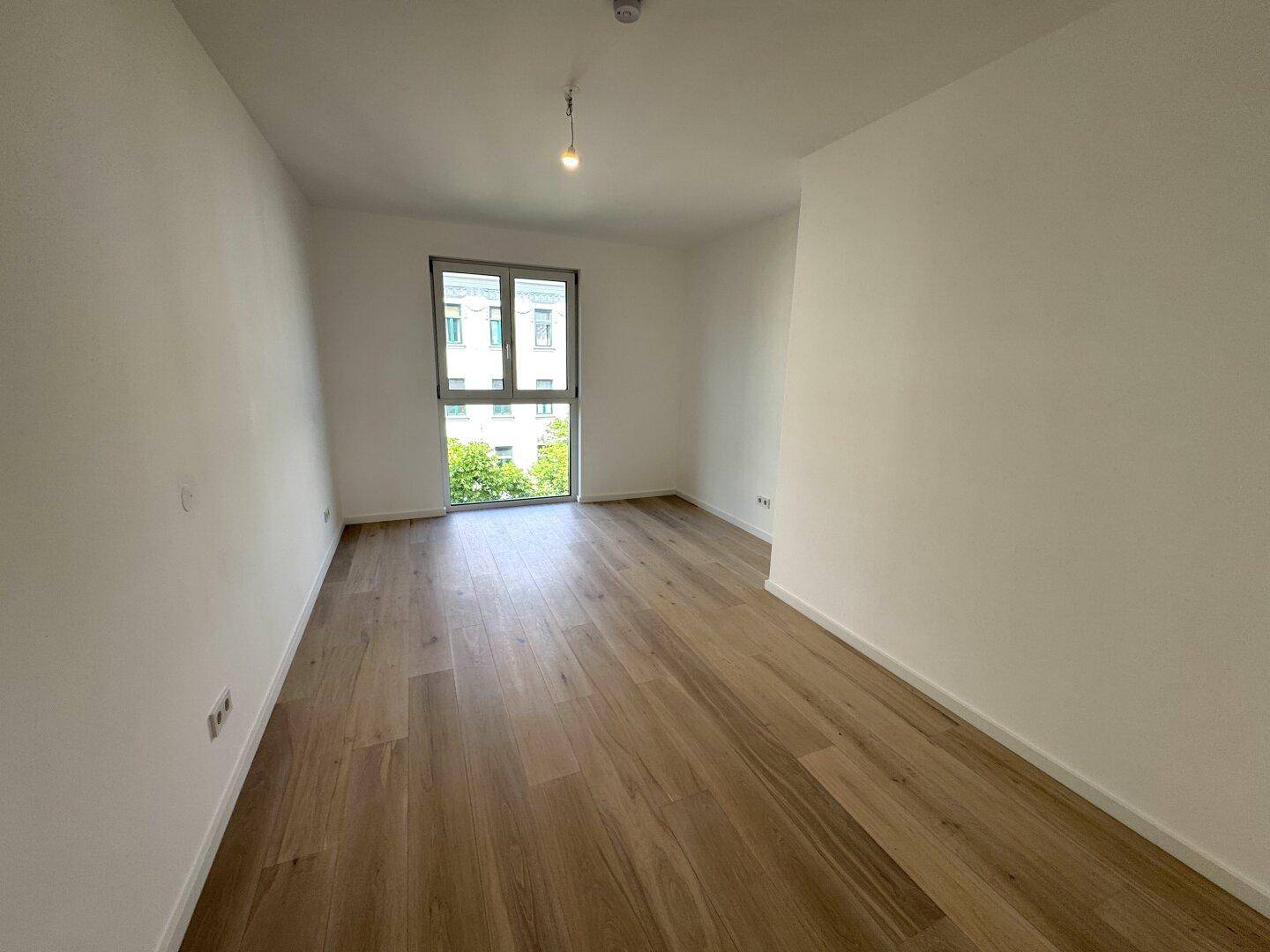 Geräumiges, helles Zimmer mit Holzboden und großem Fenster, das viel Tageslicht hereinlässt.