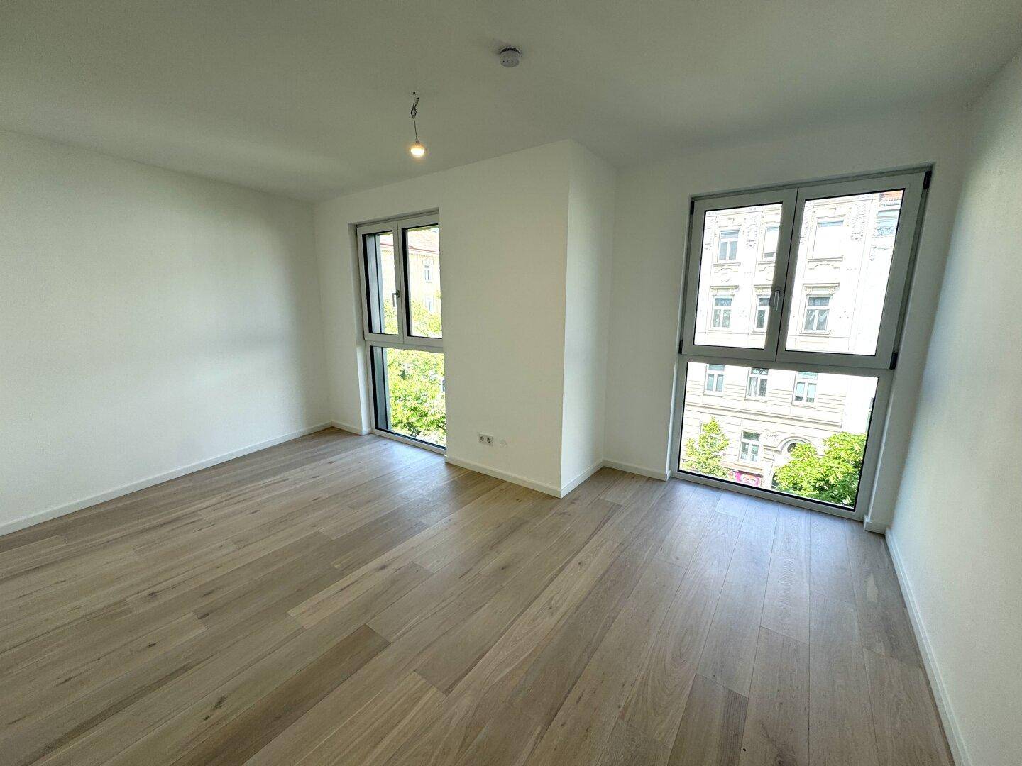 Helles, leeres Zimmer mit modernem Holzboden und großen Fenstern mit Blick ins Grüne.