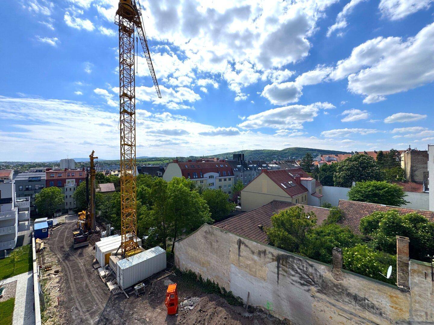 Weitläufiger Blick über die Baustelle und die umliegende Stadtlandschaft mit Hügeln im Hintergrund.