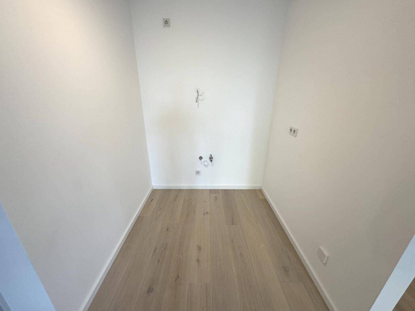 Leerer Küchenraum mit hellem Holzfußboden und vorbereiteten Anschlüssen für eine Einbauküche.