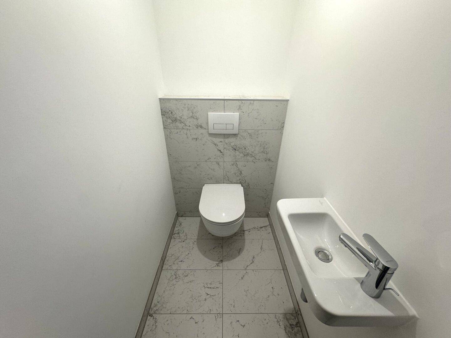 Modernes Gäste-WC mit wandhängender Toilette und kleinem Waschbecken, gefliest in Marmoroptik.