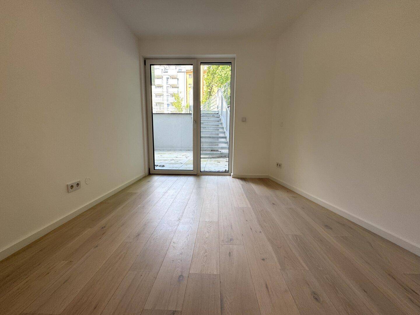 Helles, leeres Zimmer mit hellem Holzparkett und Zugang zu einer Außenterrasse über eine Glastür.
