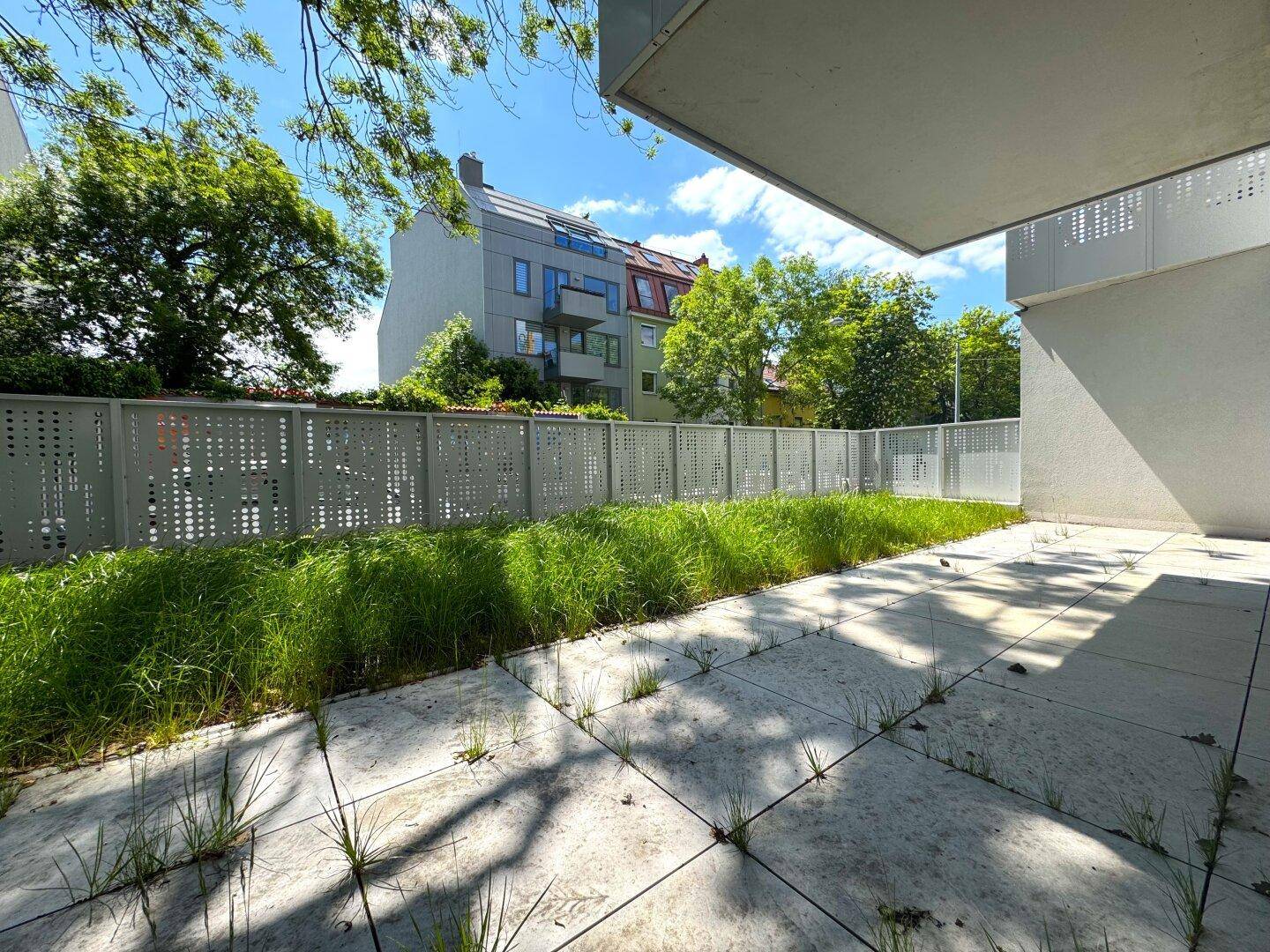 Außenbereich mit gepflasterter Terrasse und Rasenstreifen, umgeben von einer modernen Zaunanlage.