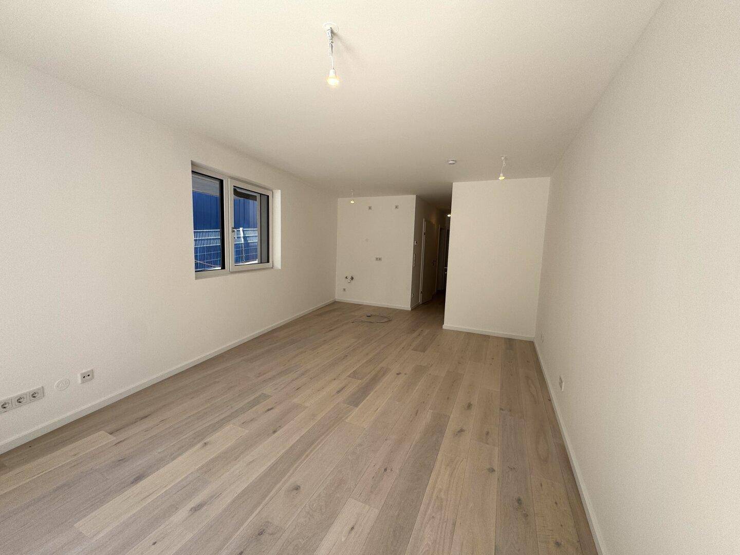 Leerer Raum mit hellem Holzboden und einem Fenster, das viel Tageslicht hereinlässt.