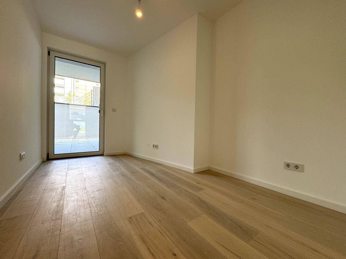Heller, leerer Raum mit Holzboden und Zugang zu einem Außenbereich durch eine Glastür.