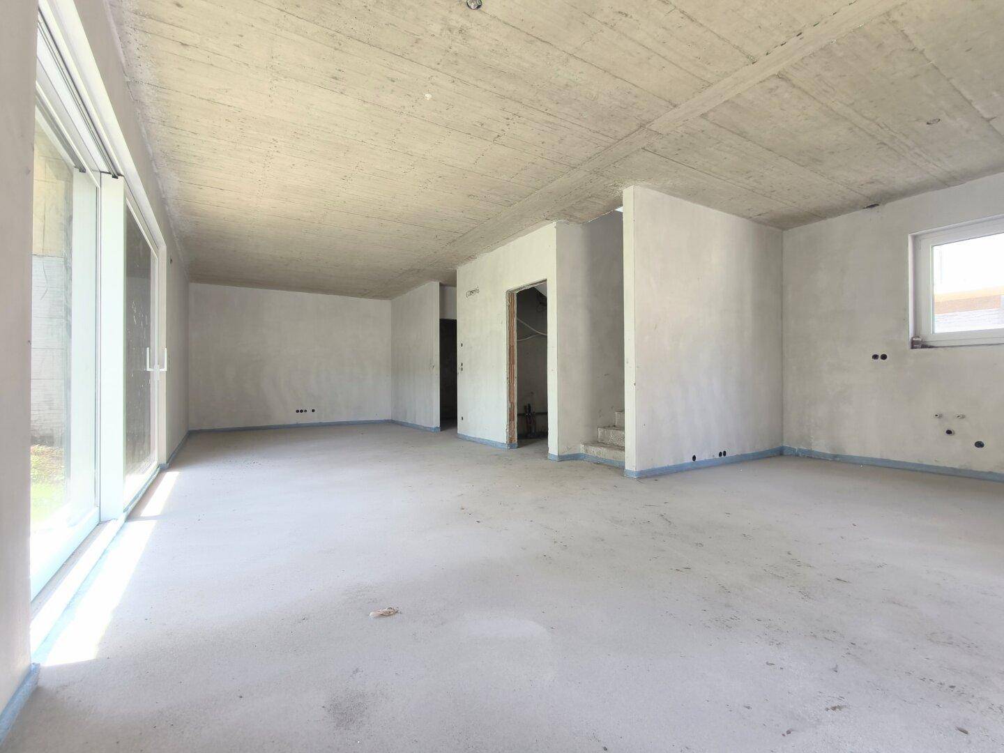 Unfertiger Wohnraum mit großen Schiebetüren und rohem Betonboden, bereit für den Ausbau.