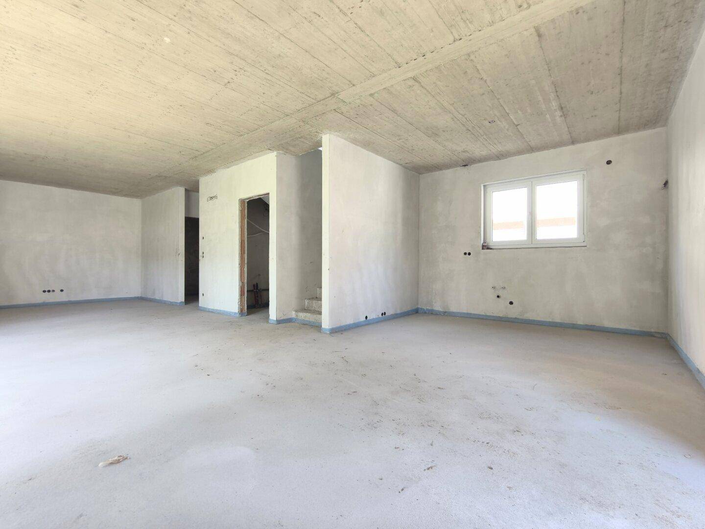 Geräumiger, unfertiger Innenraum mit Betonboden und einem Fenster, das Tageslicht hereinlässt.