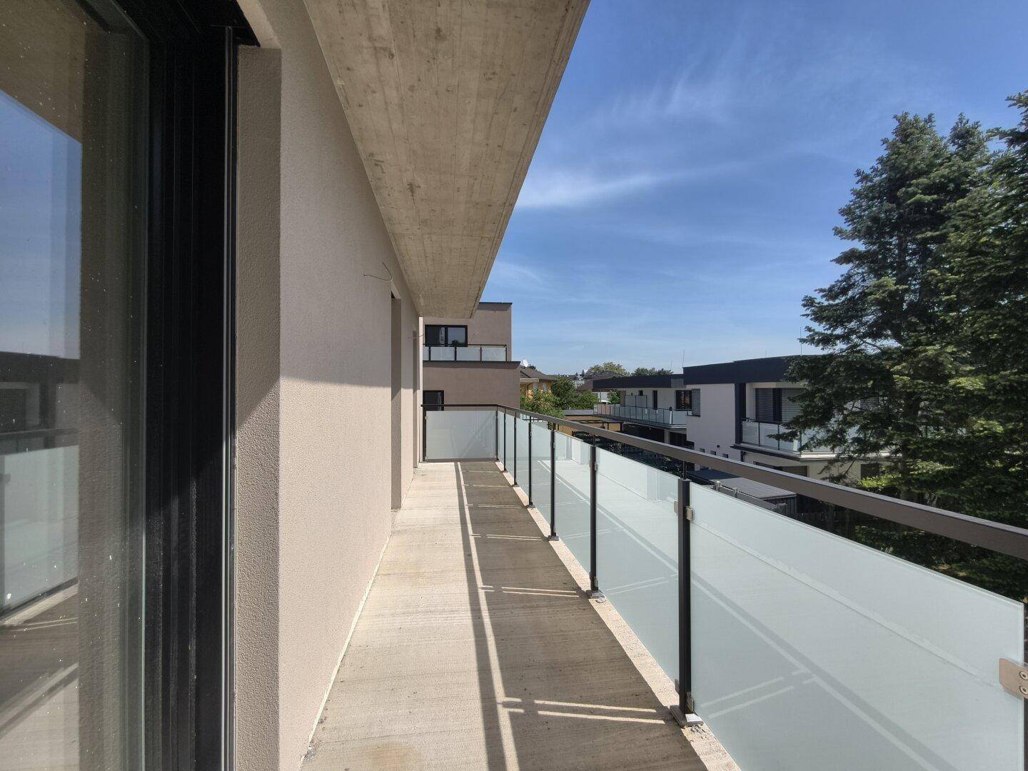Langer Balkon mit Glasgeländer und Blick auf die umliegenden Gebäude und grüne Bäume.