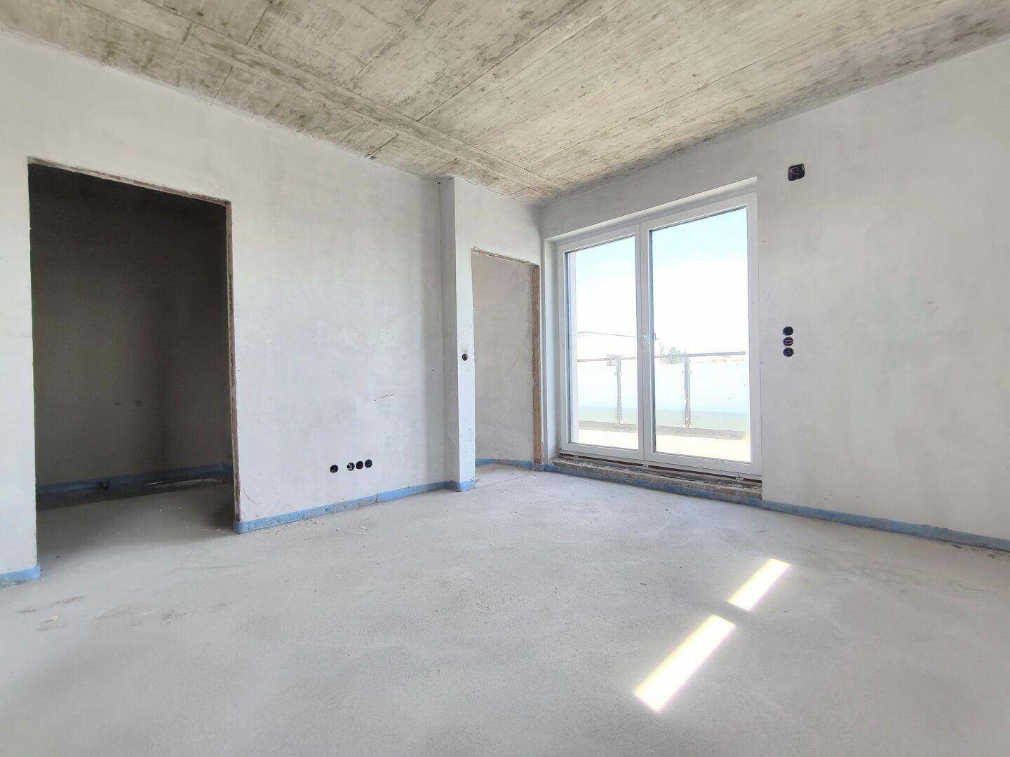 Geräumiger, unfertiger Raum mit großen Schiebetüren und Betonboden, ideal für einen Wohnbereich.