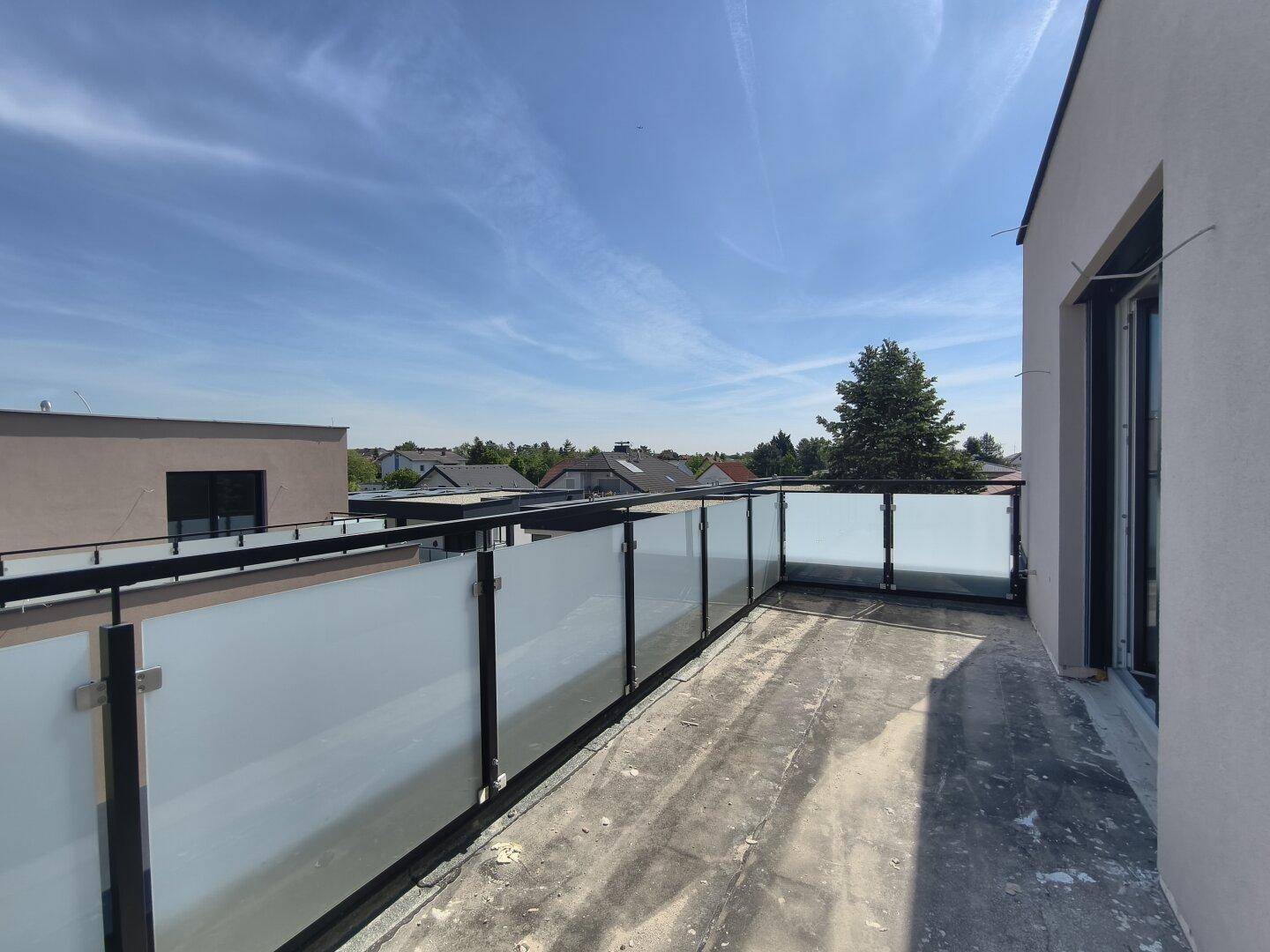 Unfertige Dachterrasse mit Glasgeländer und weitem Blick über die Dächer der Nachbarschaft.