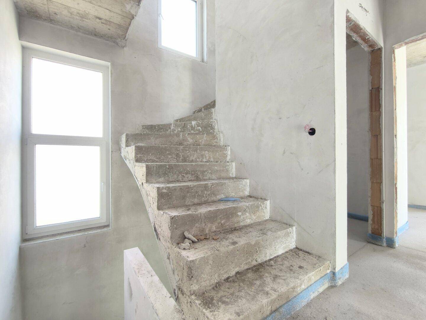 Unfertiges Treppenhaus mit Betonstufen und großen Fenstern, die den Raum erhellen.