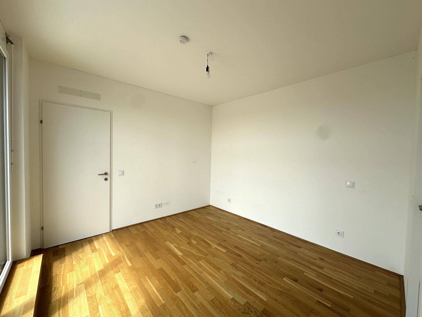 Helles Schlafzimmer mit Parkettboden, weißen Wänden und Zugang zu einem weiteren Raum.
