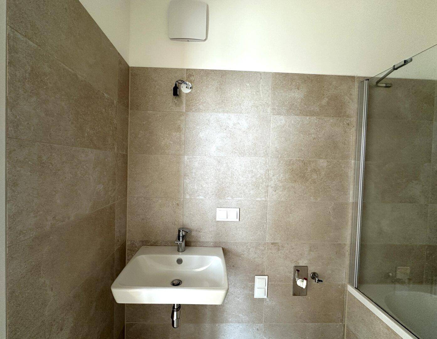 Modernes Badezimmer mit Waschbecken, Badewanne und beigefarbenen Fliesen, ausgestattet mit Abluftventilator.