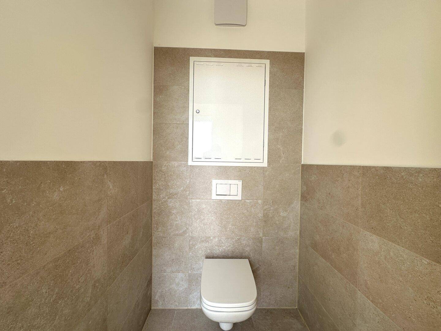 Separates WC mit Wand-WC und beigefarbenen Fliesen, ausgestattet mit Abluftventilator.