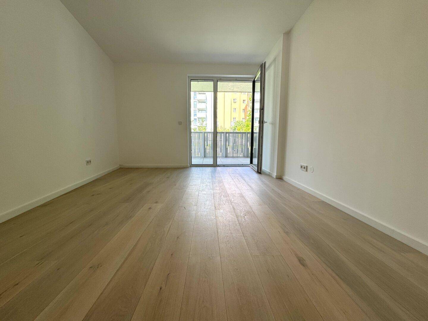 Leerer Raum mit hellem Holzparkett und großen Fenstern, die viel Tageslicht hereinlassen.