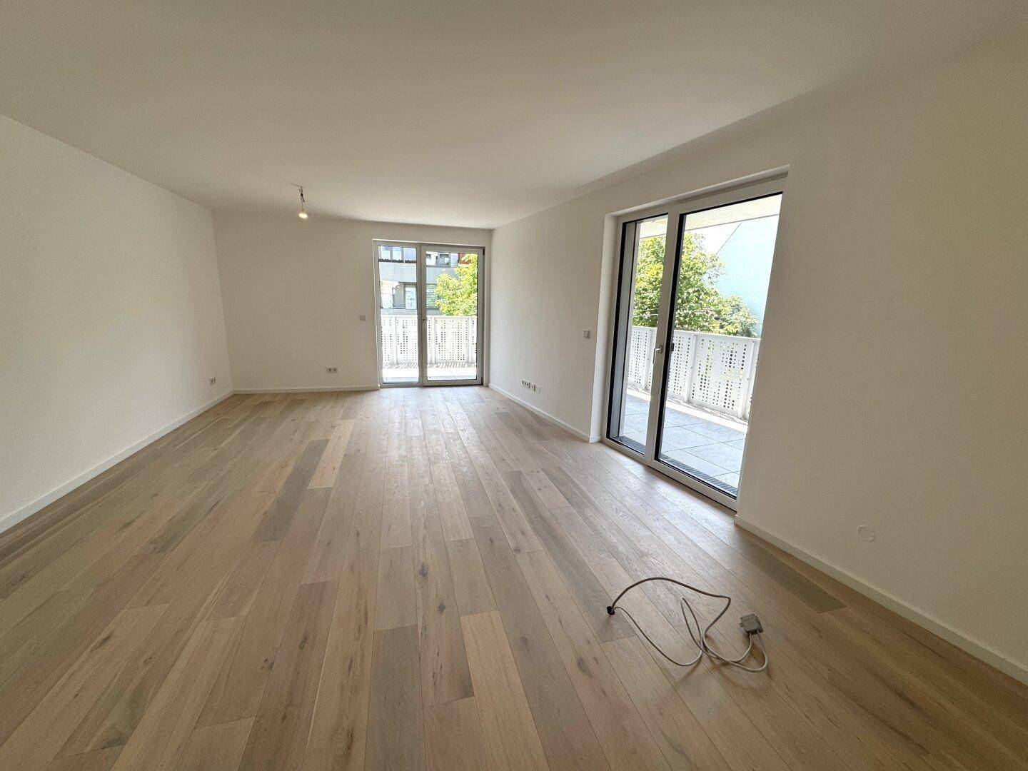 Geräumiger, heller Raum mit Holzplankenboden und zwei großen Fenstertüren, die zu einem Balkon führen.