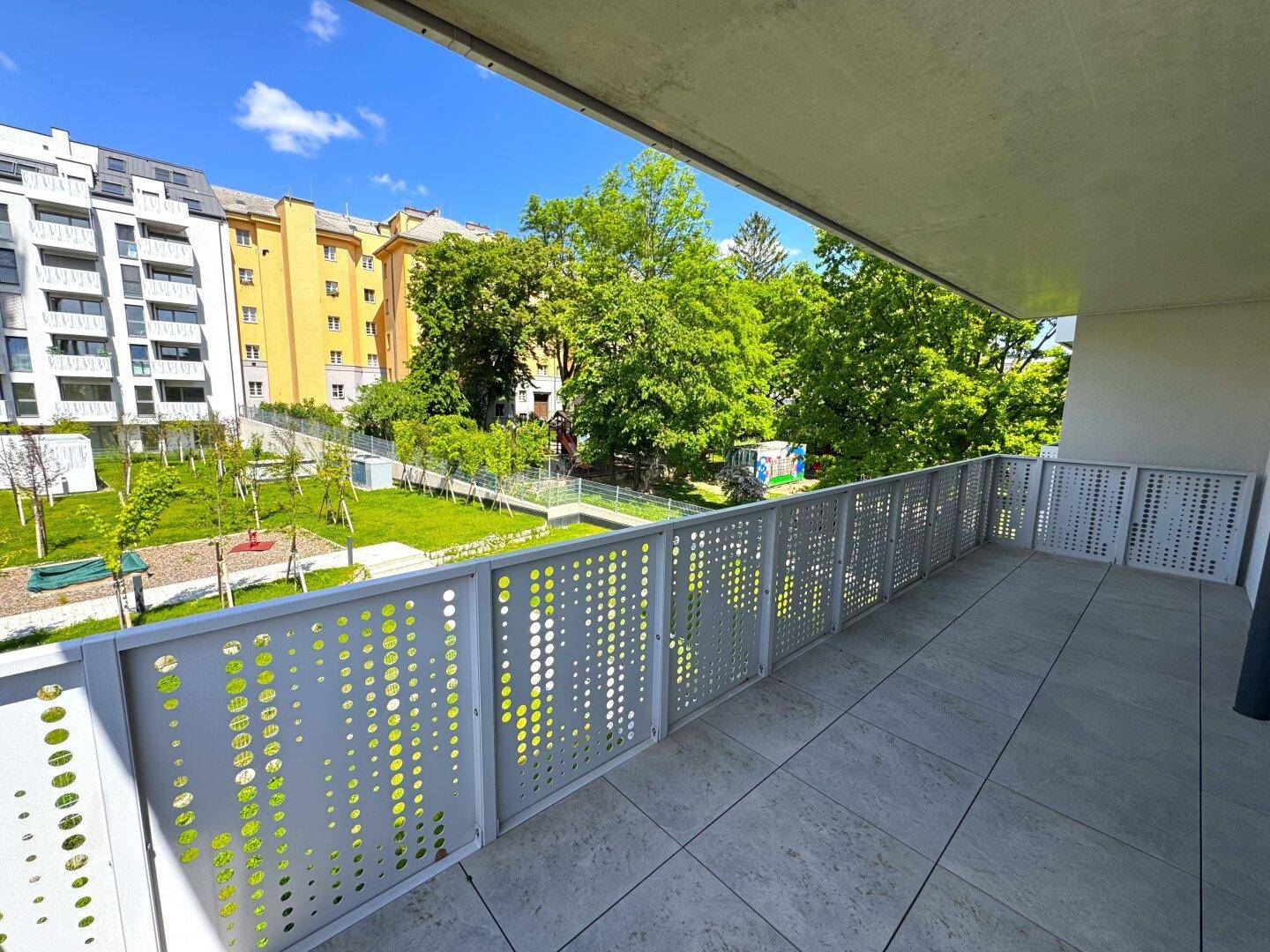Sonniger Balkon mit moderner Lochblechbrüstung und Blick auf die grüne Gartenanlage und umliegende Gebäude.