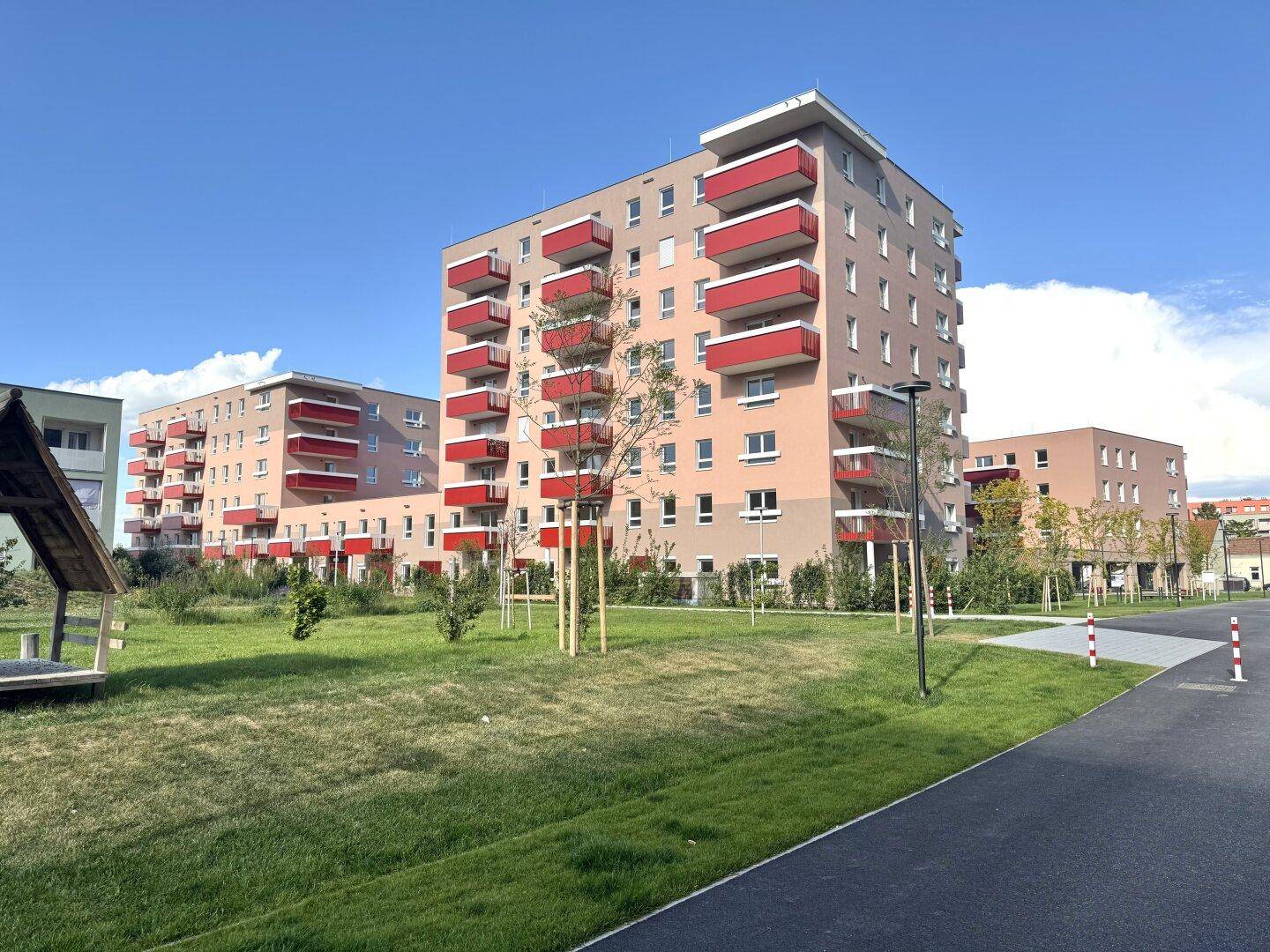 Moderne Wohngebäude mit roten Balkonen und gepflegten Grünflächen unter blauem Himmel.