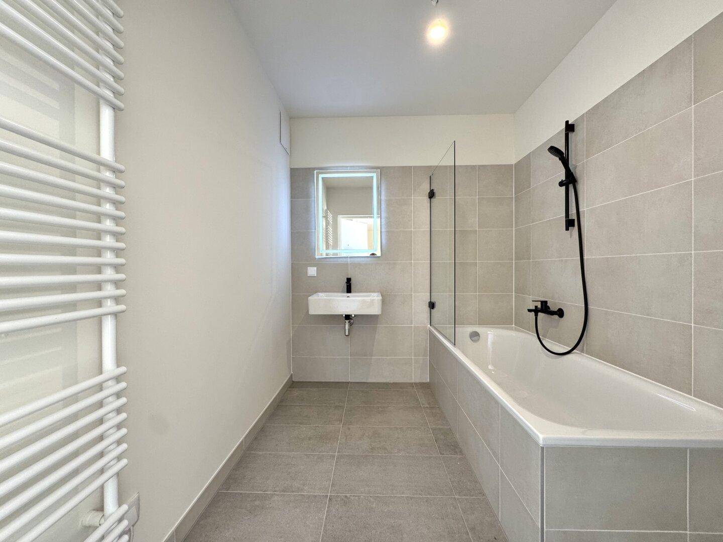 Modernes Badezimmer mit grauen Fliesen, Badewanne mit Duschwand und beheiztem Handtuchhalter.