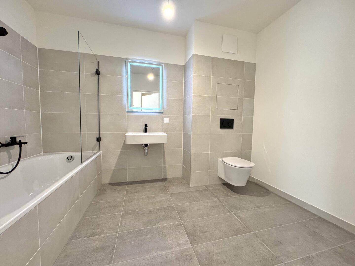 Modernes Badezimmer mit grauen Fliesen, Badewanne, wandhängendem WC und kleinem Waschbecken.