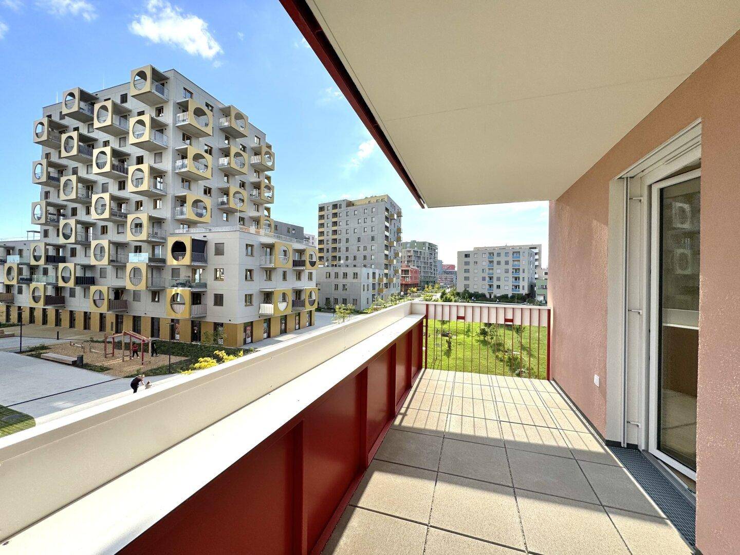 Heller Balkon mit rotem Geländer und Blick auf markante moderne Wohngebäude und Grünflächen.