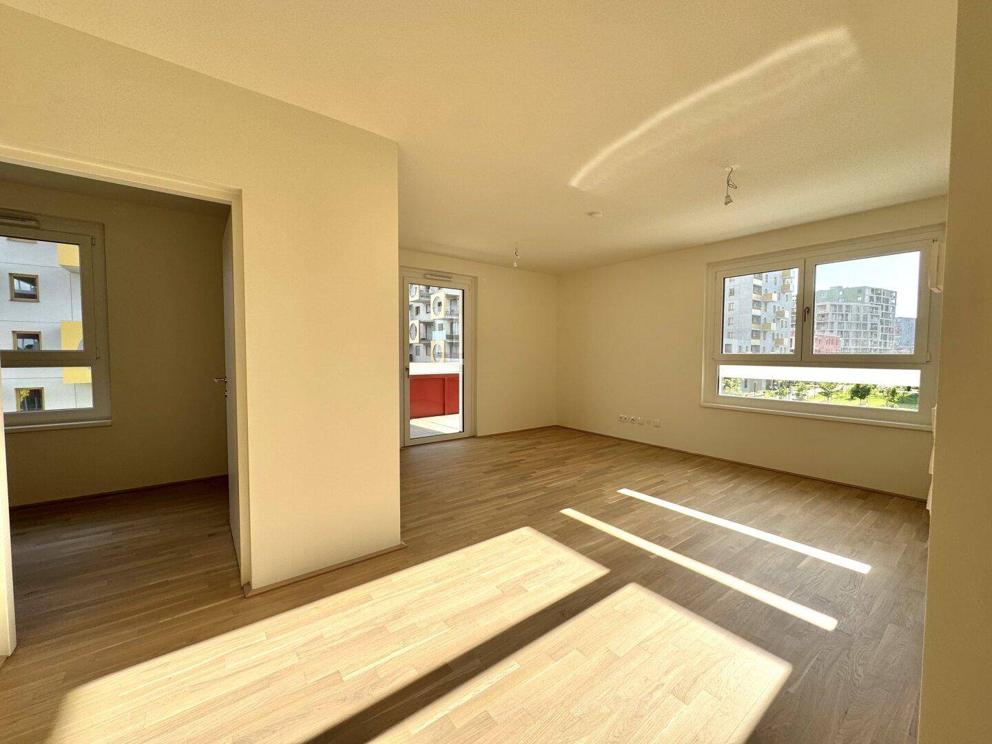 Lichtdurchfluteter, leerer Wohnraum mit hellem Laminatboden und großen Fenstern.