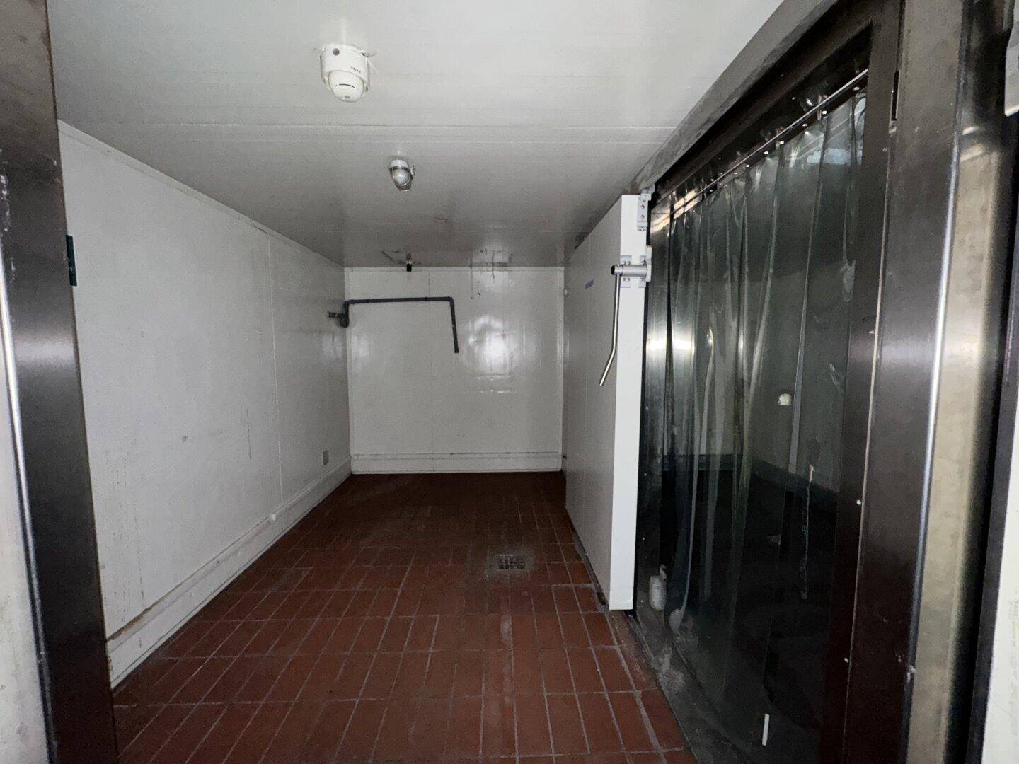 Kühlraum mit weißen Wänden, dunklem Fliesenboden und einem Lamellenvorhang am Eingang.