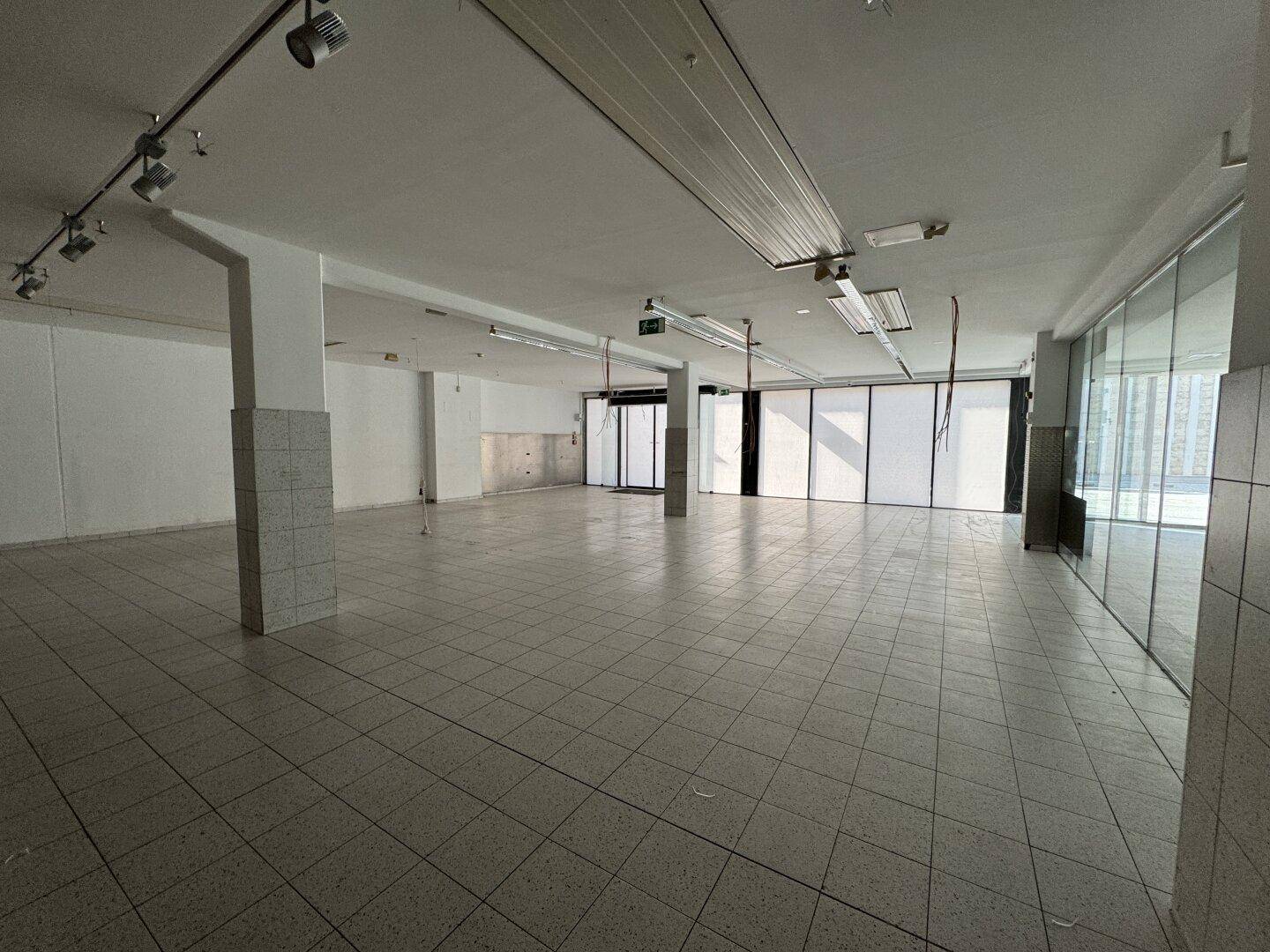 Geräumiger Gewerberaum mit Fliesenboden, Säulen, Schienenbeleuchtung und großen Fenstern.