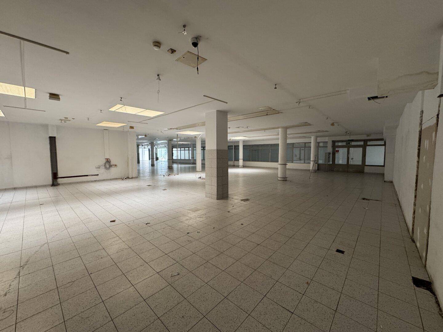 Weitläufiger, leerer Gewerberaum mit Fliesenboden, vielen Säulen und großen Fenstern im Hintergrund.