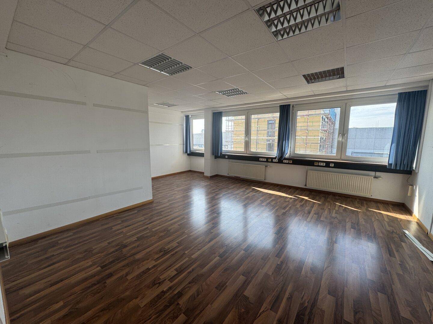 Helles Büro mit Holzboden, Rasterdecke und mehreren Fenstern, bietet viel Tageslicht.