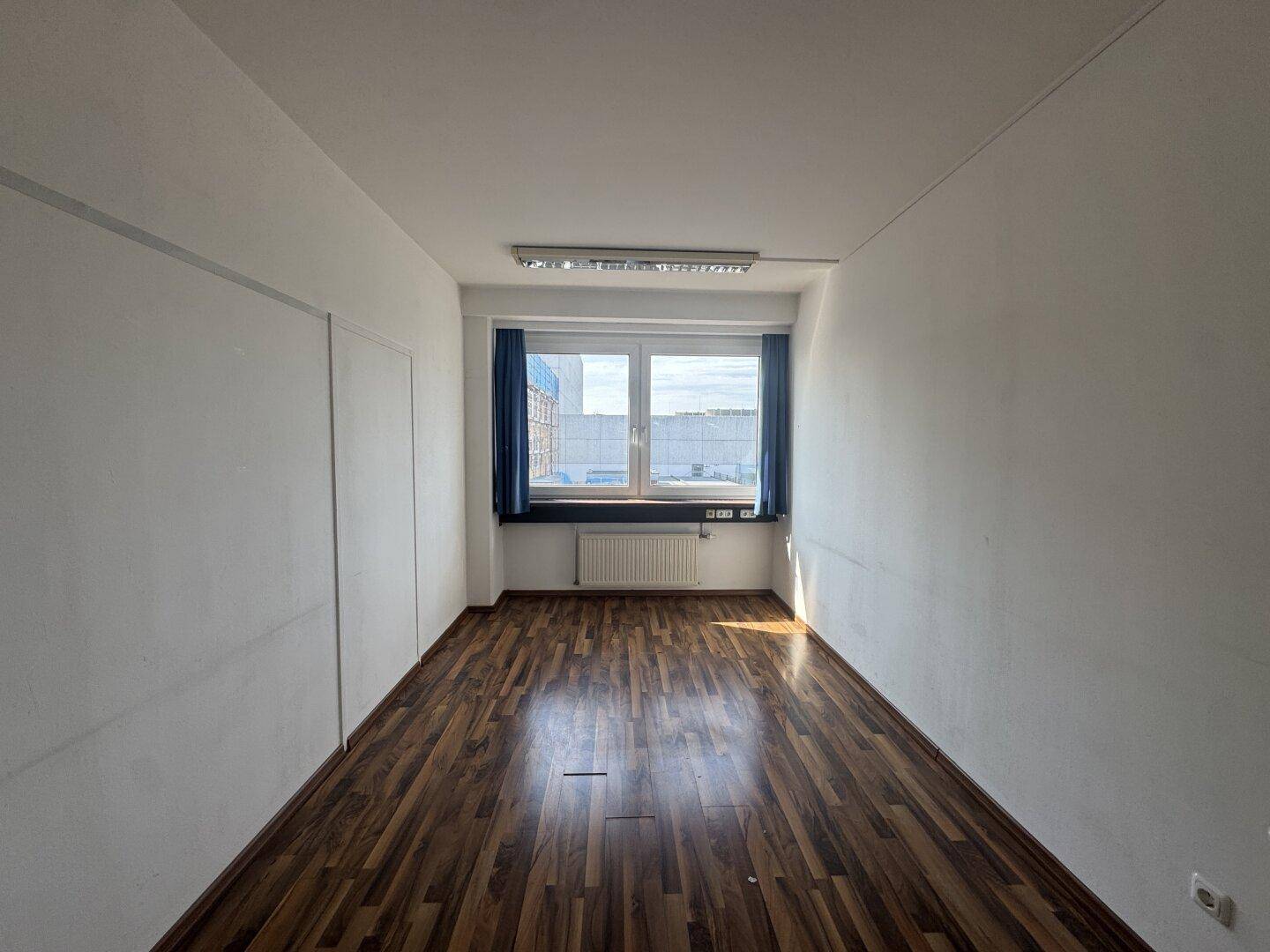Schmaler Büroraum mit Holzboden, Fenster und Deckenbeleuchtung, geeignet für Einzelbüros.