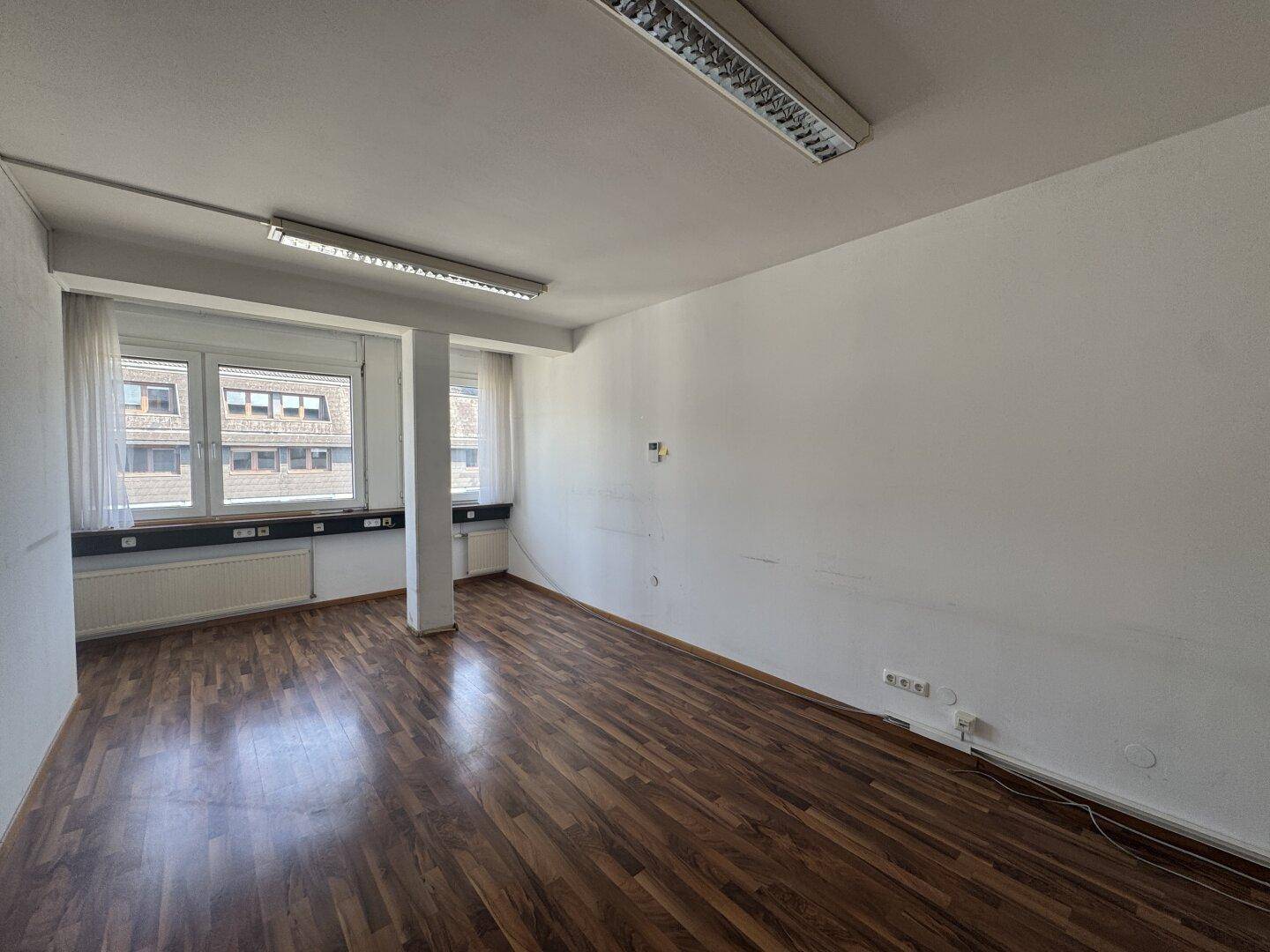 Helles Büro mit Holzboden, großen Fenstern und Deckenbeleuchtung, bietet viel Tageslicht.