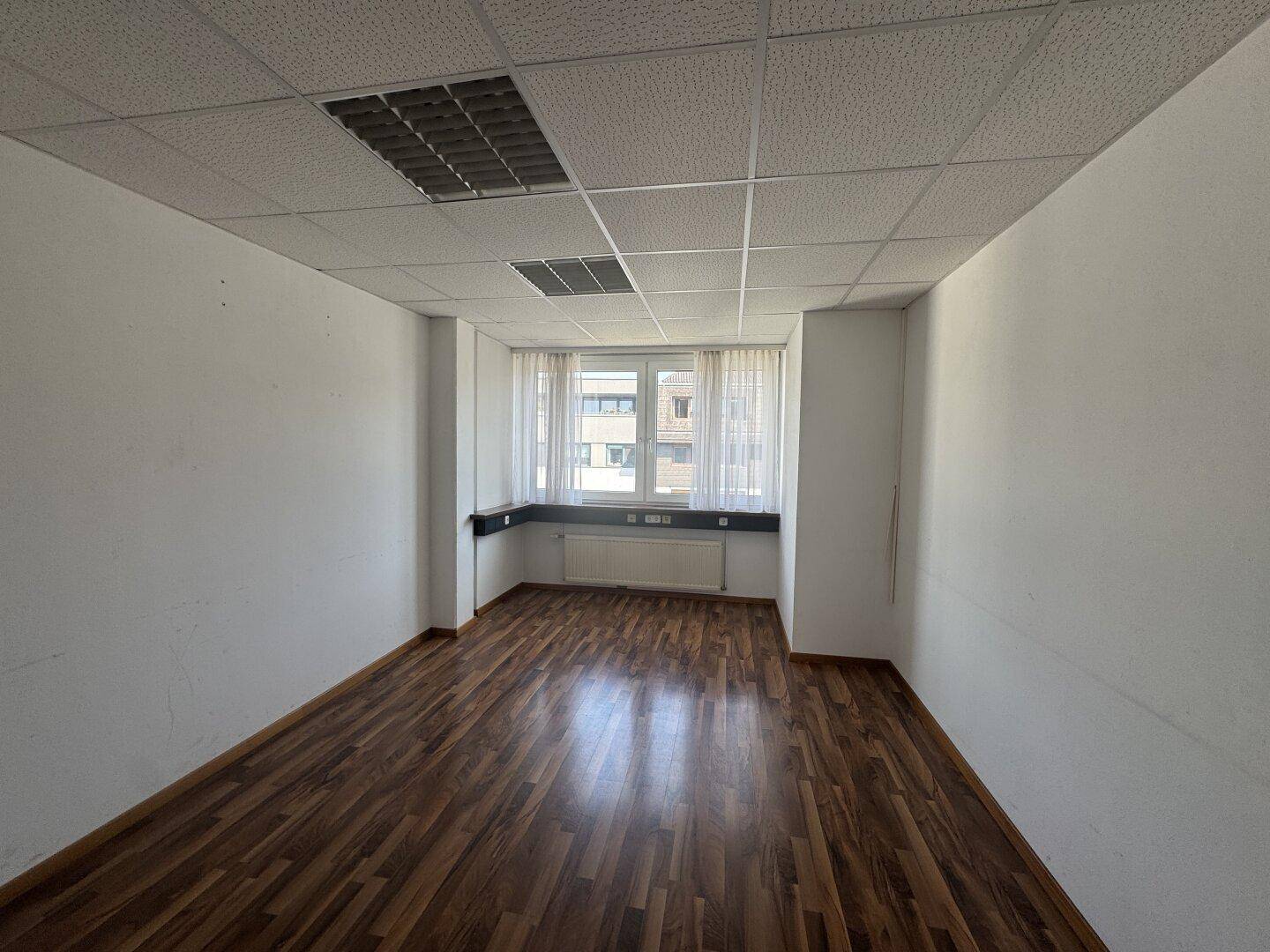 Helles Büro mit Holzboden, Rasterdecke und Fenster mit Blick nach draußen.