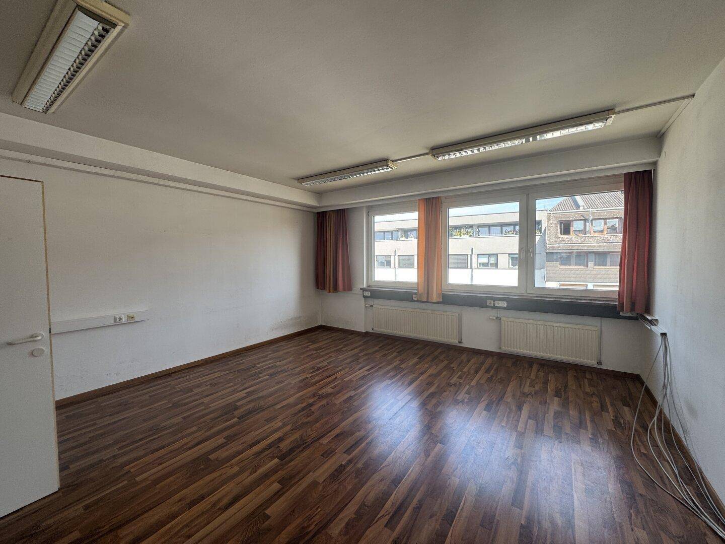 Geräumiges Büro mit Holzboden, großen Fenstern und Deckenbeleuchtung, bietet viel Tageslicht.