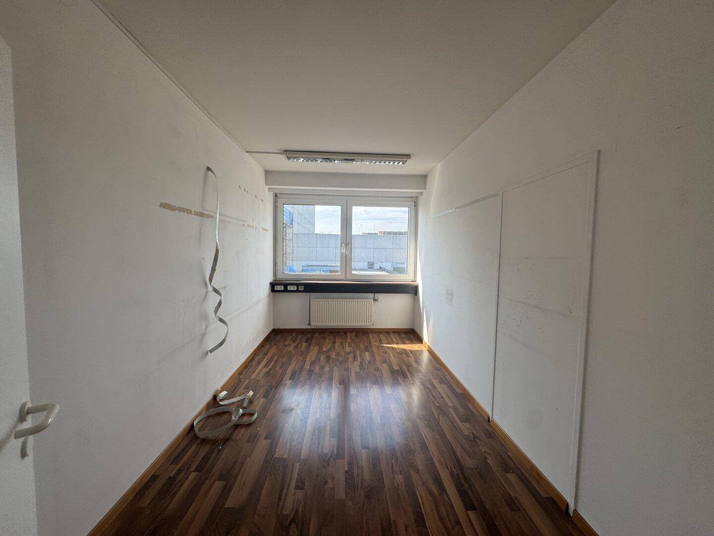 Schmaler Büroraum mit Holzboden, Fenster und Deckenbeleuchtung, ideal für Einzelarbeitsplätze.