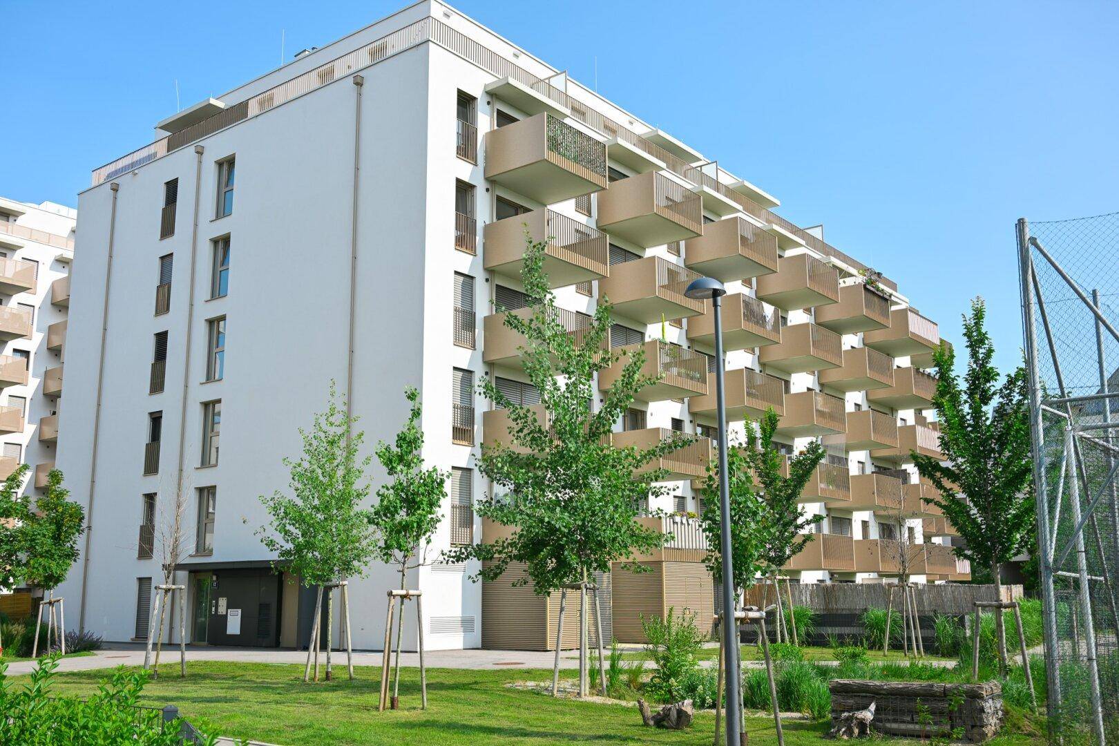 Modernes Mehrfamilienhaus mit zahlreichen Balkonen, umgeben von Grünflächen und jungen Bäumen.