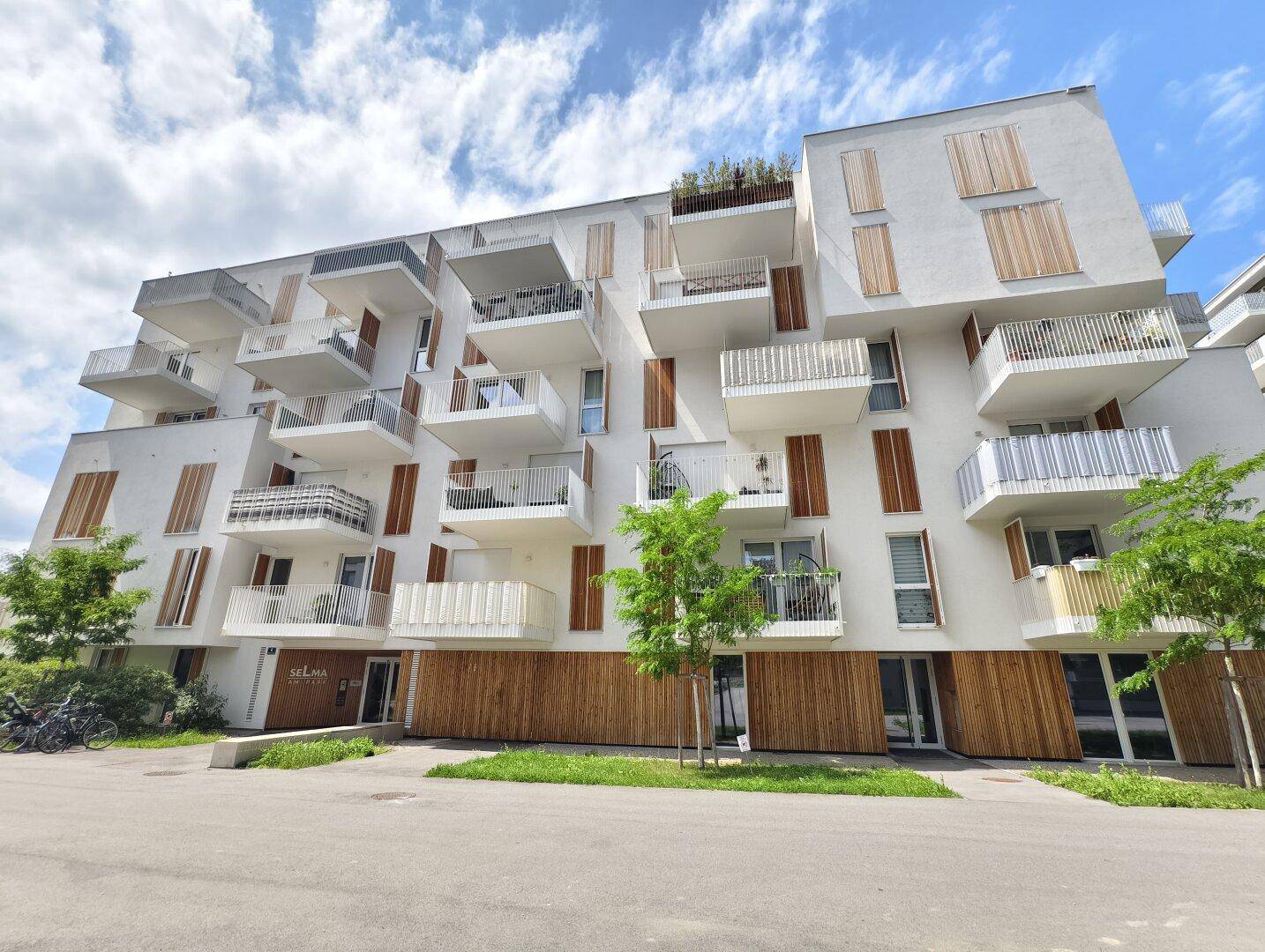 Moderne Wohnanlage mit weißer Fassade, zahlreichen Balkonen und Holzelementen unter blauem Himmel.