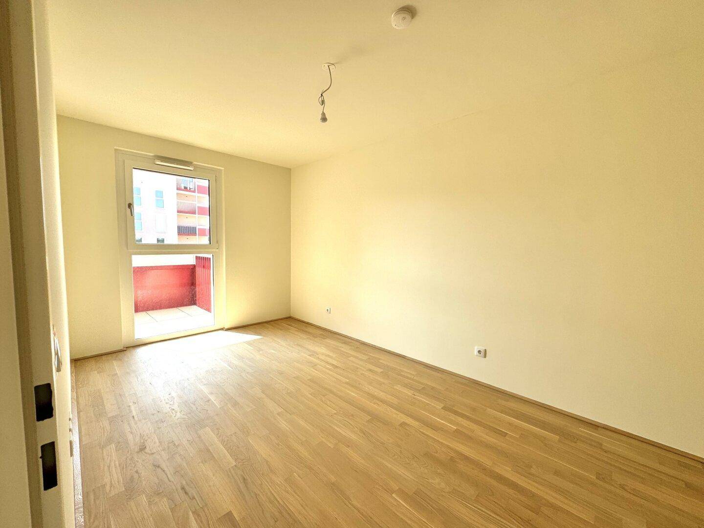 Leeres Schlafzimmer mit hellem Parkettboden und Fenster mit Blick auf die Umgebung.