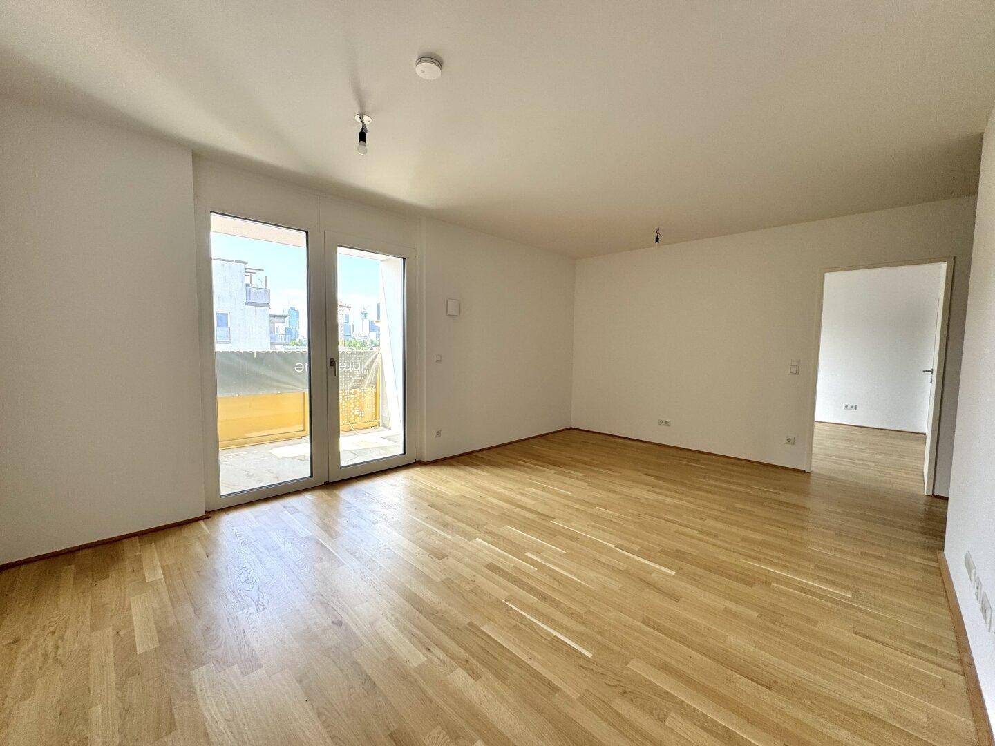 Helles, leeres Wohnzimmer mit Holzboden und großen Fenstern, die Zugang zu einem Außenbereich bieten.