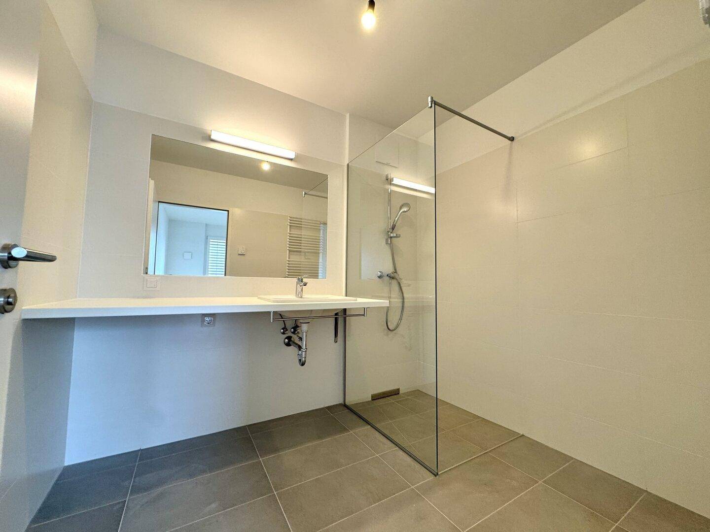 Modernes Badezimmer mit ebenerdiger Dusche, großem Spiegel und hellen Fliesen an Wänden und Boden.