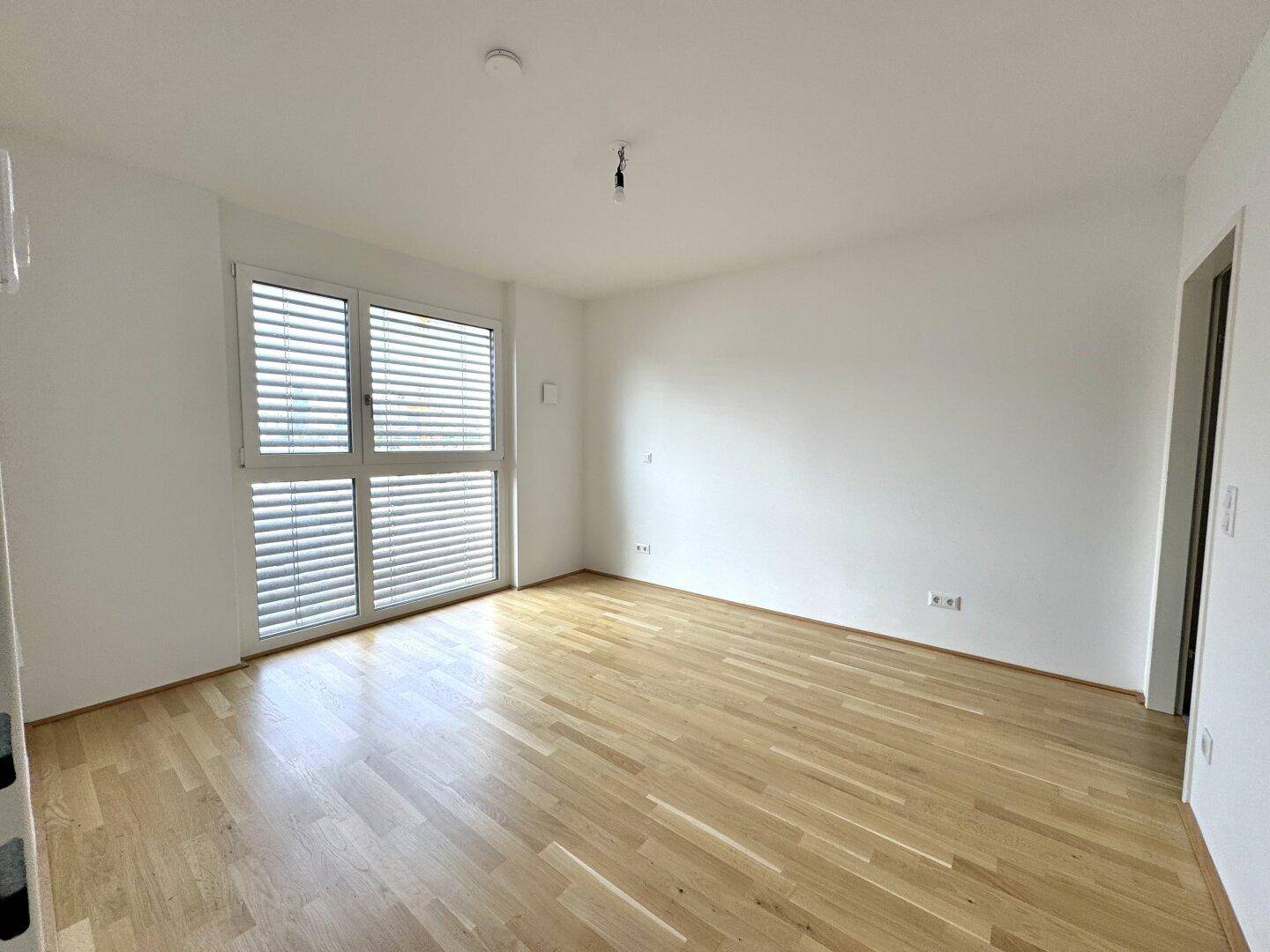 Leeres Schlafzimmer mit Holzboden und großen Fenstern, ausgestattet mit Jalousien für Privatsphäre.