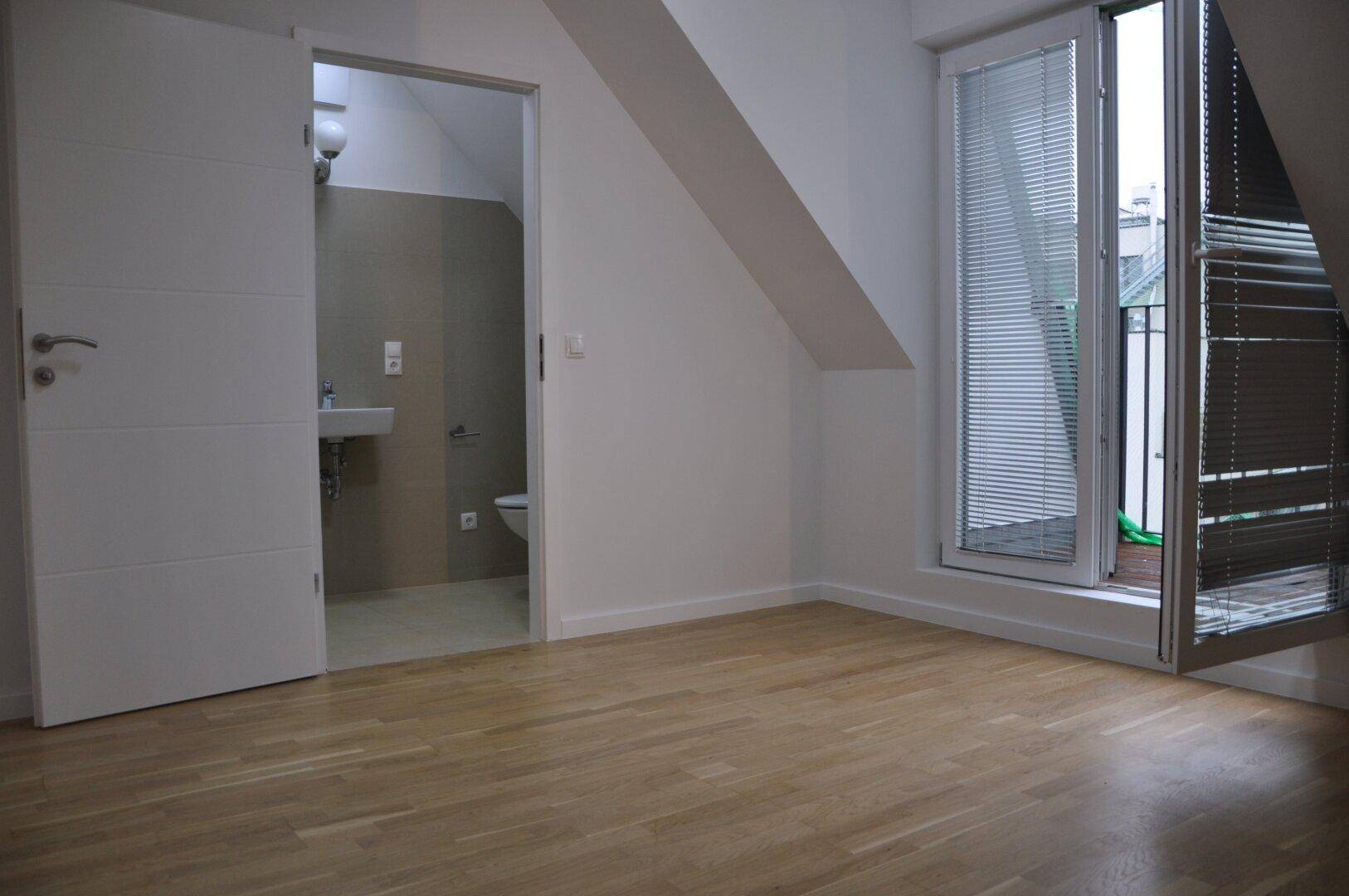 Helles Schlafzimmer mit Holzfußboden, Dachschrägen und Zugang zu einem Badezimmer und Balkon.