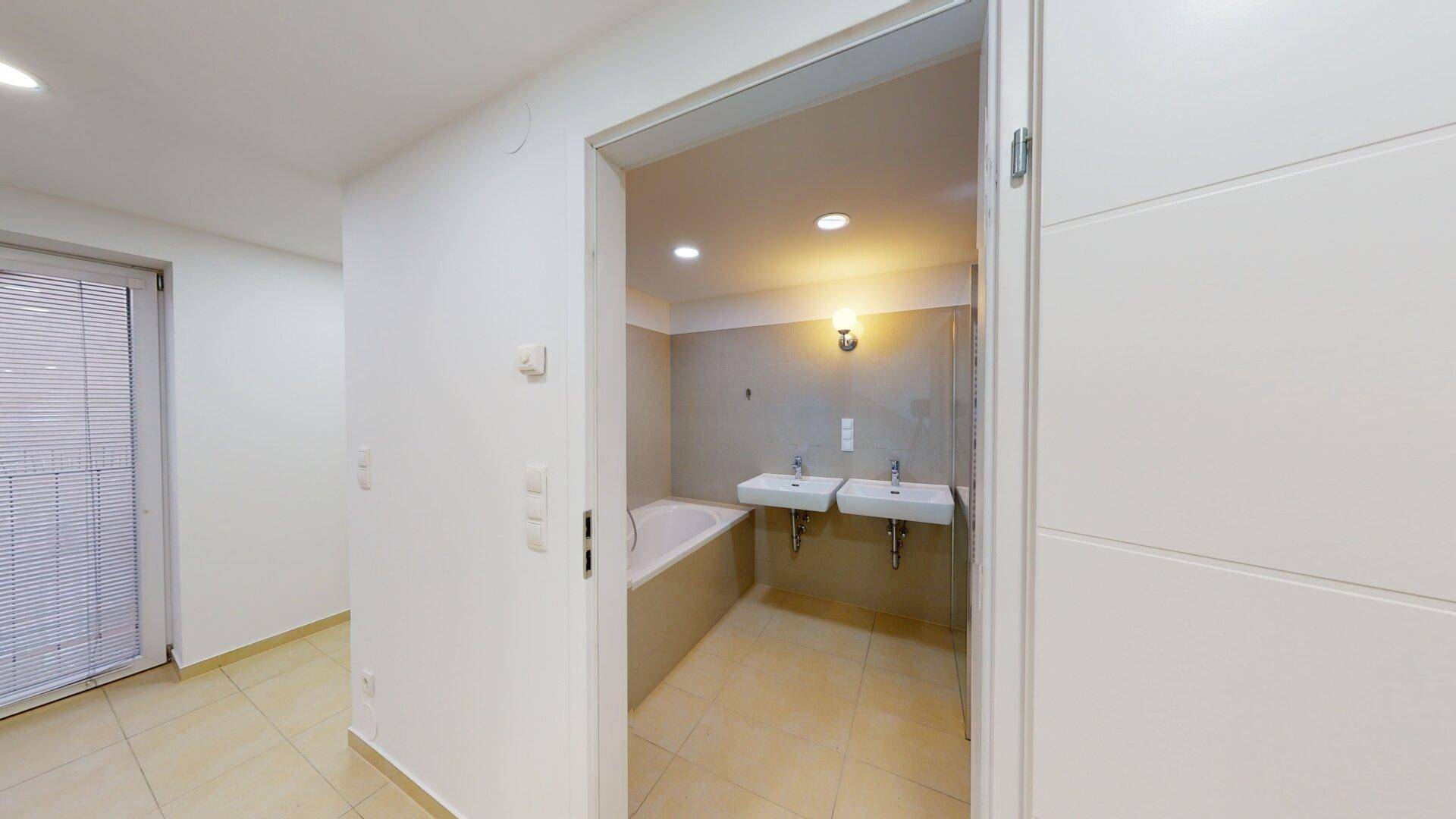 Modernes Badezimmer mit Badewanne, Doppelwaschbecken und hellen Fliesen, vom Flur aus sichtbar.
