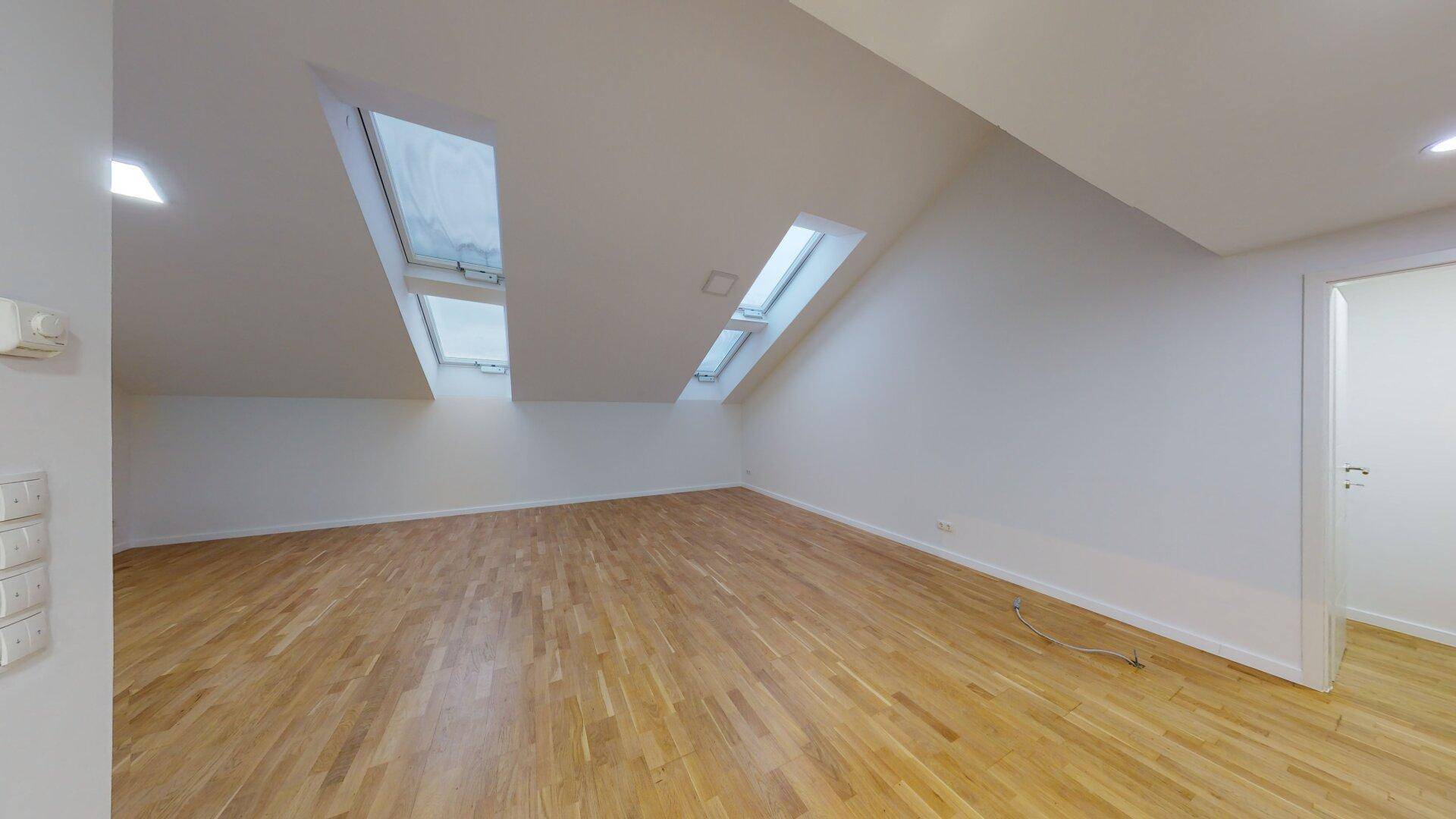 Geräumiger Dachgeschossraum mit Holzfußboden, schrägen Wänden und mehreren Dachfenstern.