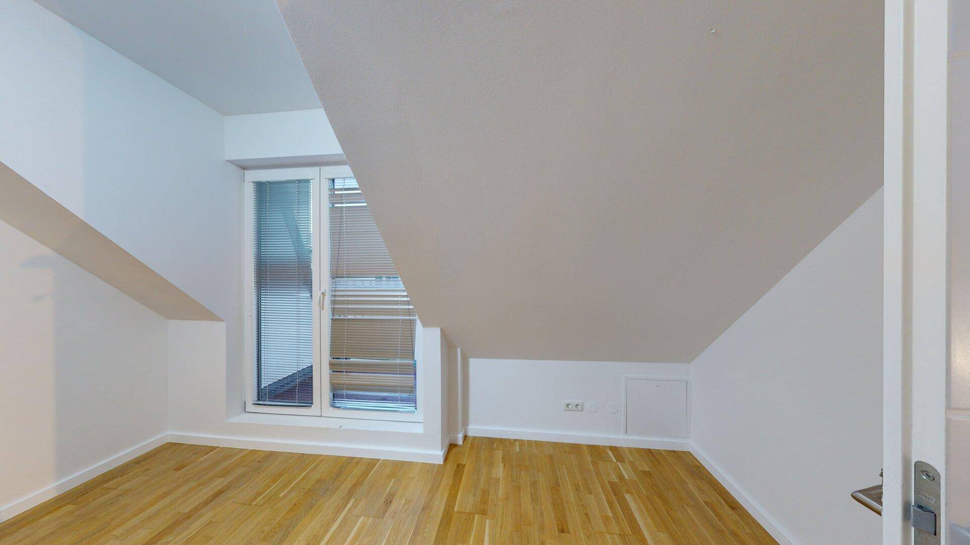 Helles Zimmer mit Holzfußboden, Dachschrägen und einem Fenster mit Jalousien.