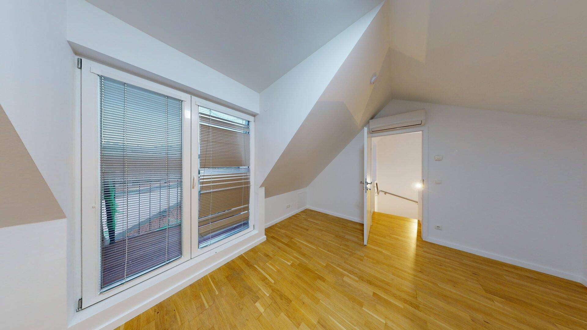 Helles Zimmer mit Holzfußboden, Dachschrägen und einem Fenster mit Jalousien.